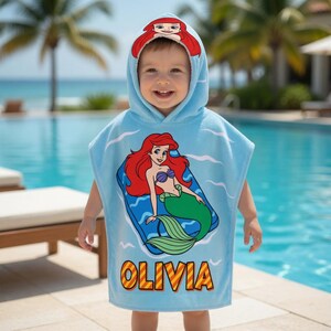 K&ouml;nnte beinhalten: Ein hellblaues Strandtuch mit Kapuze und einem Cartoon-Bild von Arielle, der kleinen Meerjungfrau. Das Handtuch tr&auml;gt den Namen "Olivia" in Gelb und Orange. Die Kapuze hat ein Design von Arielle mit rotem Haar.
