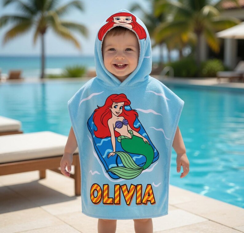 K&ouml;nnte beinhalten: Ein hellblaues Strandtuch mit Kapuze und einem Cartoon-Bild von Arielle, der kleinen Meerjungfrau. Das Handtuch tr&auml;gt den Namen "Olivia" in Gelb und Orange. Die Kapuze hat ein Design von Arielle mit rotem Haar.