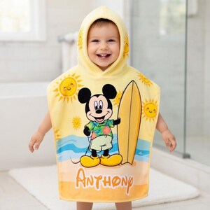 Puede incluir: Toalla de playa con capucha amarilla con Mickey Mouse sosteniendo una tabla de surf. La toalla tiene un dise&ntilde;o de escena de playa con soles sonrientes y el nombre "Anthony" impreso. Dise&ntilde;ado para ni&ntilde;os.