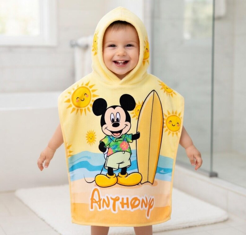 Puede incluir: Toalla de playa con capucha amarilla con Mickey Mouse sosteniendo una tabla de surf. La toalla tiene un dise&ntilde;o de escena de playa con soles sonrientes y el nombre "Anthony" impreso. Dise&ntilde;ado para ni&ntilde;os.
