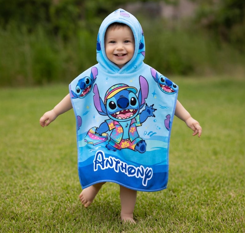 Puede incluir: Una toalla de playa con capucha azul claro con un personaje de dibujos animados, Stitch, con una camisa hawaiana. La toalla tiene un patr&oacute;n de olas azules y blancas y el nombre "Anthony" en escritura blanca. El ni&ntilde;o lleva la toalla y sonr&iacute;e.