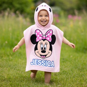 May include: A pink hooded towel featuring a Minnie Mouse design with a polka dot bow and the name "JESSICA" in blue. The towel is worn by a child in a grassy outdoor setting.