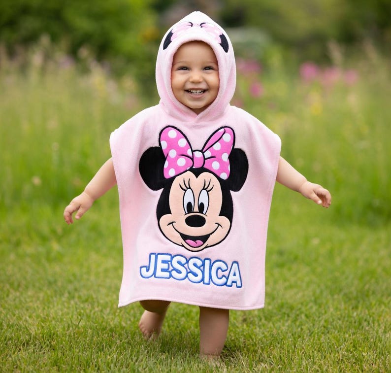 May include: A pink hooded towel featuring a Minnie Mouse design with a polka dot bow and the name "JESSICA" in blue. The towel is worn by a child in a grassy outdoor setting.