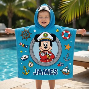 Op de afbeelding: Een lichtblauwe strandhanddoek met capuchon met een cartoon Mickey Mouse kapitein ontwerp. De handdoek heeft nautische thema graphics, waaronder een scheepsstuur, ankers en een zeilboot. De naam "JAMES" is op de handdoek gedrukt.