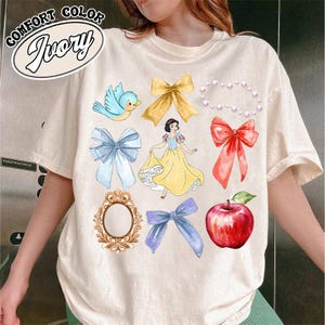 K&ouml;nnte beinhalten: Ein wei&szlig;es T-Shirt mit einem Aufdruck von Schneewittchen, einem blauen Vogel, einem roten Apfel, einem Spiegel und mehreren Schleifen in Rot, Blau und Gelb. Der Text "Comfort Color Ivory" ist in der oberen linken Ecke des Shirts gedruckt.