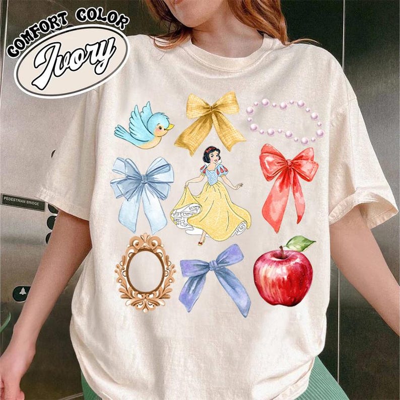 K&ouml;nnte beinhalten: Ein wei&szlig;es T-Shirt mit einem Aufdruck von Schneewittchen, einem blauen Vogel, einem roten Apfel, einem Spiegel und mehreren Schleifen in Rot, Blau und Gelb. Der Text "Comfort Color Ivory" ist in der oberen linken Ecke des Shirts gedruckt.
