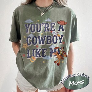 May include: A moss green short-sleeve t-shirt featuring the text "YOU'RE A COWBOY LIKE ME" in dark blue letters. The graphic includes a brown cowboy hat, a rocket ship, a yellow star, a colorful ball, and a cartoon cowboy riding a brown horse.