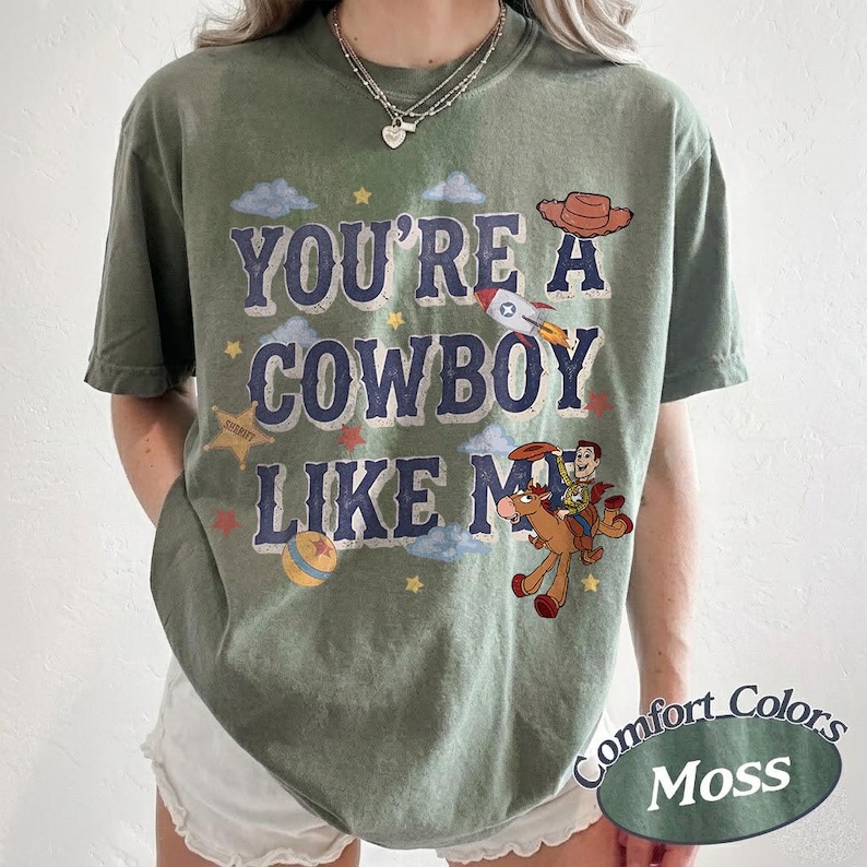 May include: A moss green short-sleeve t-shirt featuring the text "YOU'RE A COWBOY LIKE ME" in dark blue letters. The graphic includes a brown cowboy hat, a rocket ship, a yellow star, a colorful ball, and a cartoon cowboy riding a brown horse.