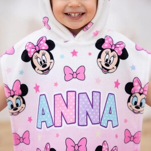 May include: A hooded child's towel featuring Minnie Mouse. The towel is white with pink and blue accents, and has multiple Minnie Mouse faces, pink bows, and stars. The name "ANNA" is printed in large letters across the front.
