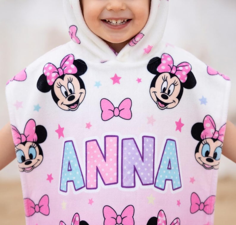 May include: A hooded child's towel featuring Minnie Mouse. The towel is white with pink and blue accents, and has multiple Minnie Mouse faces, pink bows, and stars. The name "ANNA" is printed in large letters across the front.