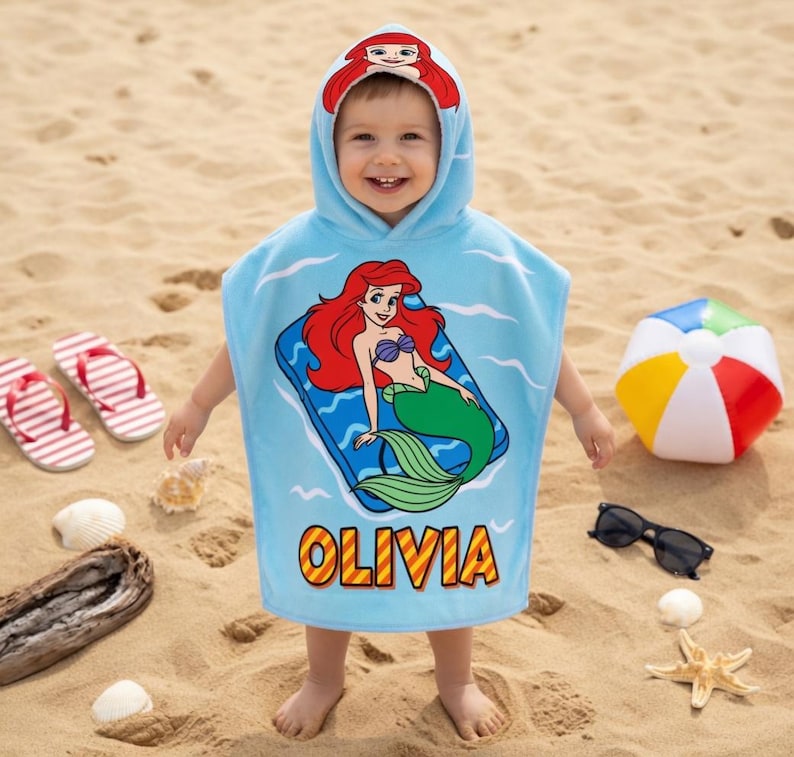 K&ouml;nnte beinhalten: Ein hellblaues Strandtuch mit Kapuze, das ein Cartoon-Meerjungfrau-Design und den Namen "OLIVIA" in Gelb zeigt. Das Handtuch hat eine Kapuze mit einem Meerjungfrau-Design. Das Bild zeigt auch einen Strandball, eine Sonnenbrille und Flip-Flops.