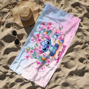 May include: A pink and blue beach towel with a cartoon Stitch character holding a tropical drink. The towel has a floral design and the name "Jessica" is written on it.