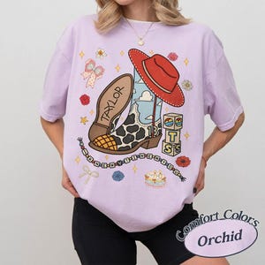 May include: A light purple t-shirt featuring a graphic print of a cow-print cowboy boot, a red hat, building blocks with the letters T, S, and 13, and a string of beads spelling I KNEW YOU WERE TROUBLE with the name TAYLOR on the boot sole.