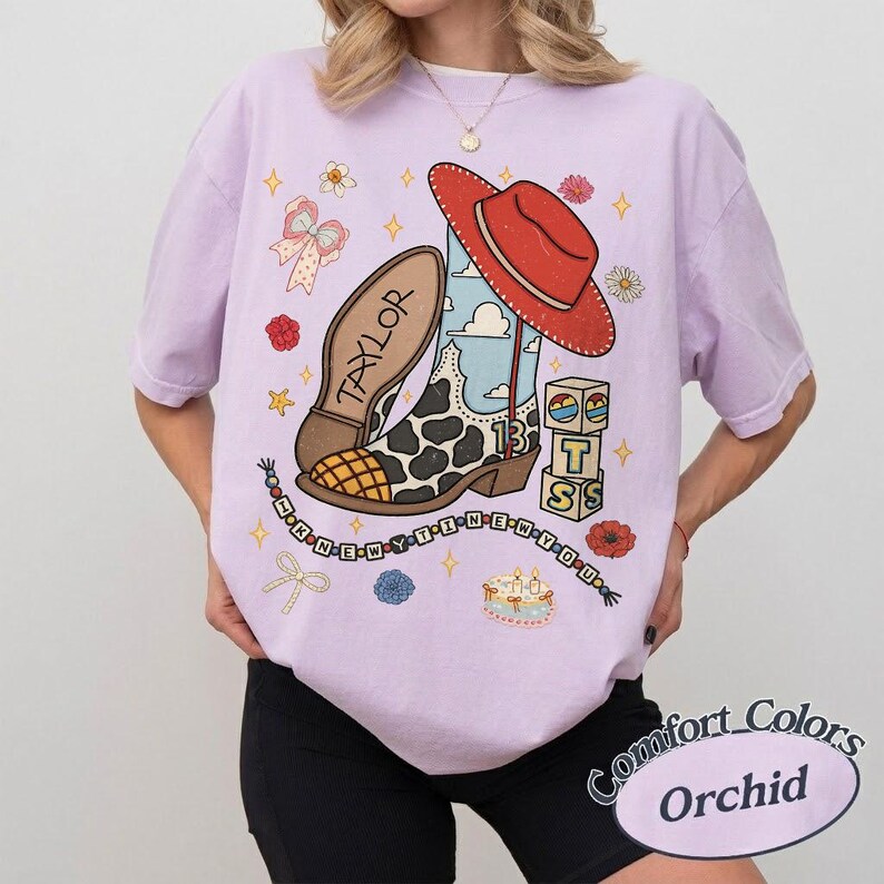 May include: A light purple t-shirt featuring a graphic print of a cow-print cowboy boot, a red hat, building blocks with the letters T, S, and 13, and a string of beads spelling I KNEW YOU WERE TROUBLE with the name TAYLOR on the boot sole.