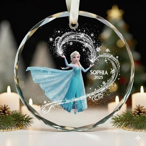 Puede incluir: Adorno navideño redondo y transparente con Elsa de Frozen con un vestido azul. El adorno tiene una cinta blanca para colgar y incluye el nombre "SOPHIA" y el año "2025" en un diseño en forma de corazón con copos de nieve.