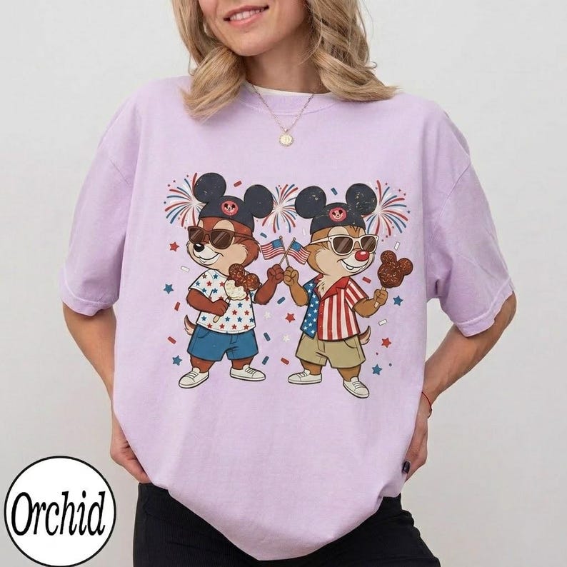 May include: A light purple t-shirt featuring a graphic of two cartoon chipmunks wearing sunglasses and patriotic outfits. They are holding American flags and treats, with fireworks and confetti in the background. The shirt has a relaxed fit.