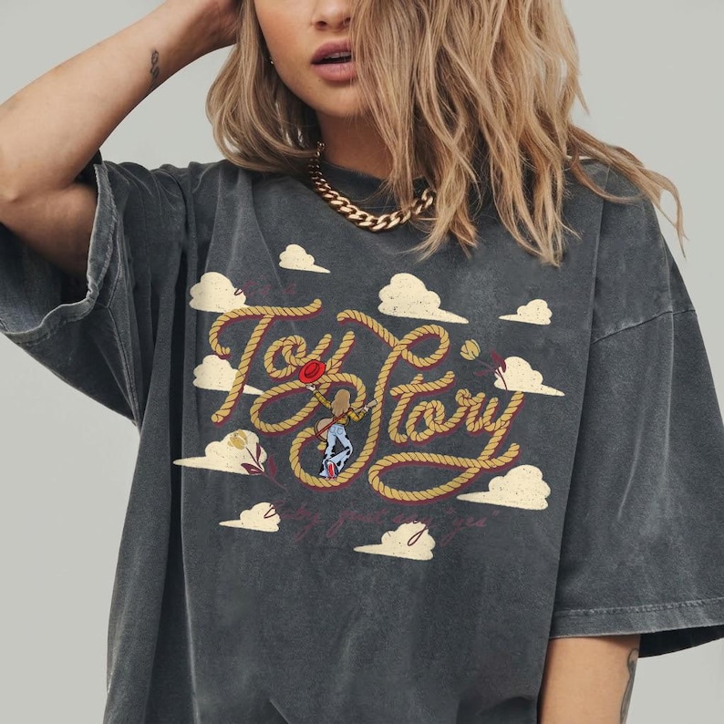 May include: A charcoal gray short-sleeve t-shirt featuring a graphic print of the text Toy Story written in a rope-like font. The design includes a small character figure, a red hat, and several beige clouds scattered across the front of the shirt.