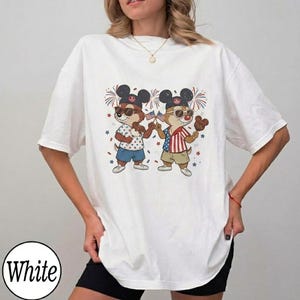 May include: White t-shirt featuring a cartoon illustration of two chipmunks wearing sunglasses and Mickey Mouse ears, holding American flags and treats. The chipmunks are dressed in patriotic outfits, with fireworks and star accents. The word "White" is printed on the bottom left.
