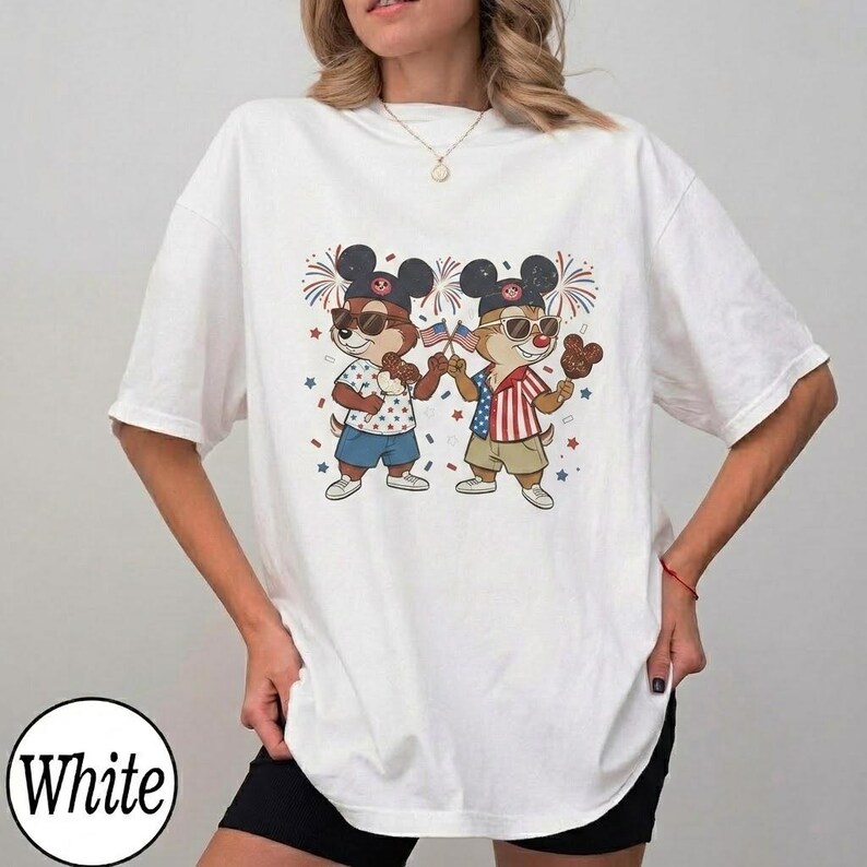 May include: White t-shirt featuring a cartoon illustration of two chipmunks wearing sunglasses and Mickey Mouse ears, holding American flags and treats. The chipmunks are dressed in patriotic outfits, with fireworks and star accents. The word "White" is printed on the bottom left.