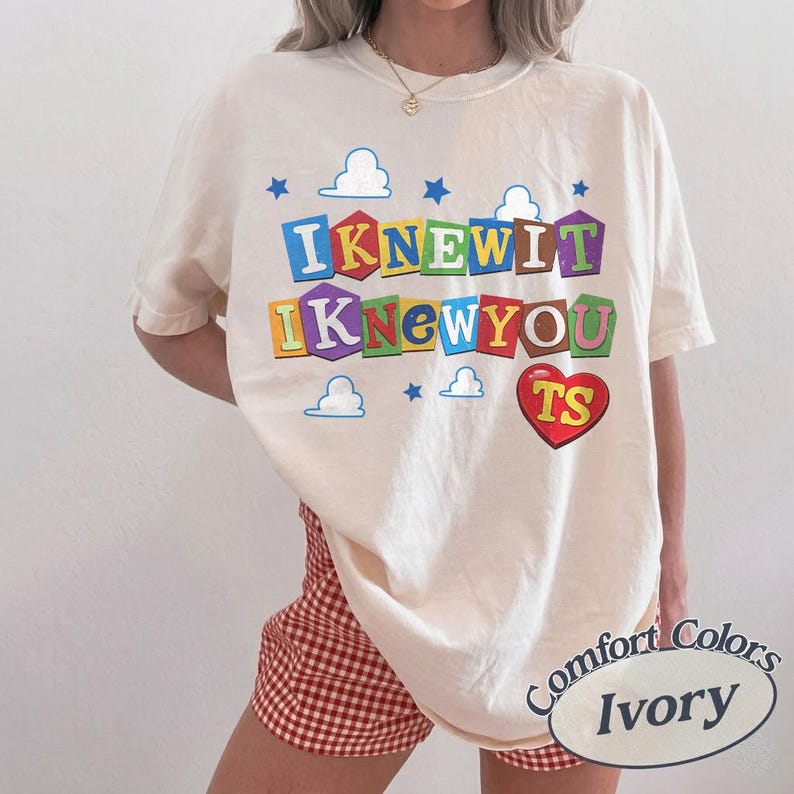 May include: An ivory short-sleeve t-shirt featuring the text "IKNEWIT IKNEWYOU" in colorful block letters on a background of blue clouds and stars. A red heart with the letters "TS" is printed below the text. A small oval label reads "Comfort Colors Ivory".