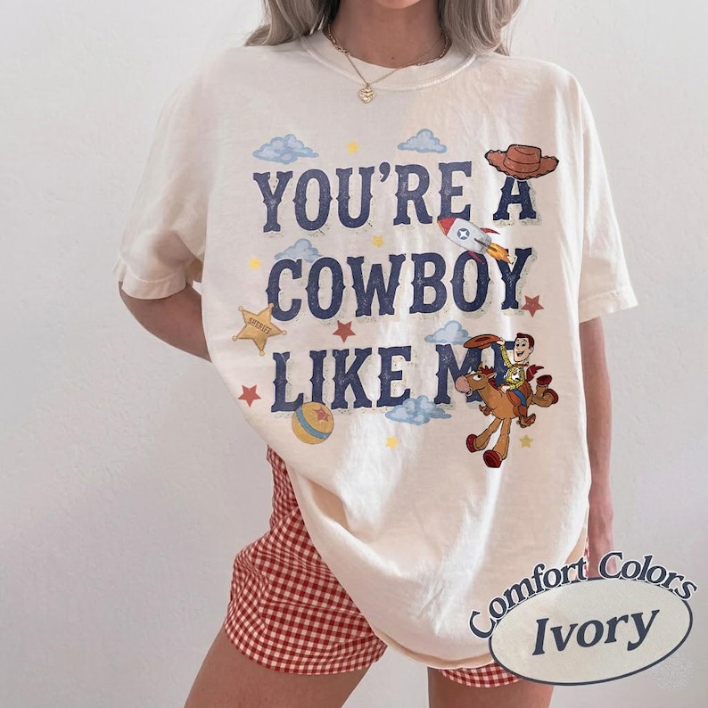 May include: An ivory short-sleeve t-shirt featuring the dark blue text "YOU'RE A COWBOY LIKE ME" alongside graphic illustrations of a cowboy on a horse, a rocket, a sheriff star, clouds, and small stars. The shirt is paired with red and white gingham shorts.