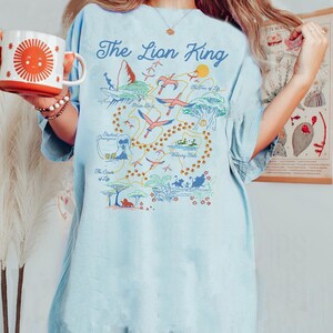 May include: A light blue t-shirt with a graphic print of a map of the Lion King movie. The map includes locations like Pride Rock, The Watering Hole, and The Tree of Life. The text "The Lion King" is printed in a bold, white font at the top of the map.