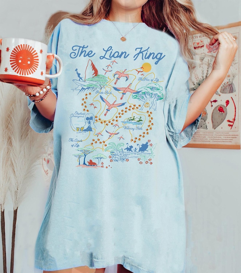 May include: A light blue t-shirt with a graphic print of a map of the Lion King movie. The map includes locations like Pride Rock, The Watering Hole, and The Tree of Life. The text "The Lion King" is printed in a bold, white font at the top of the map.