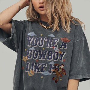 May include: A charcoal gray short-sleeve t-shirt featuring the text "YOU'RE A COWBOY LIKE ME" in blue letters. The graphic includes a brown cowboy hat, a rocket ship, a sheriff star, a colorful ball, and a cartoon character riding a brown horse.