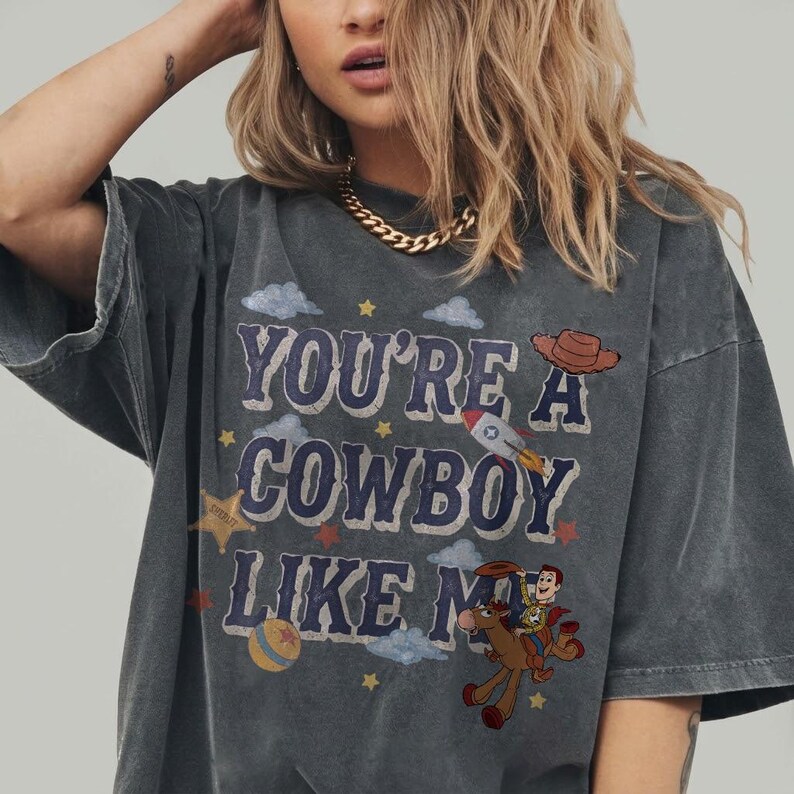 May include: A charcoal gray short-sleeve t-shirt featuring the text "YOU'RE A COWBOY LIKE ME" in blue letters. The graphic includes a brown cowboy hat, a rocket ship, a sheriff star, a colorful ball, and a cartoon character riding a brown horse.
