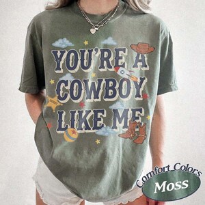 May include: A moss green short-sleeve t-shirt featuring the text YOU'RE A COWBOY LIKE ME in dark blue letters. The graphic includes a brown cowboy hat, a rocket, a yellow sheriff star, a colorful ball, and brown cowboy boots with clouds.