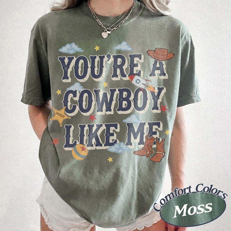 May include: A moss green short-sleeve t-shirt featuring the text YOU'RE A COWBOY LIKE ME in dark blue letters. The graphic includes a brown cowboy hat, a rocket, a yellow sheriff star, a colorful ball, and brown cowboy boots with clouds.