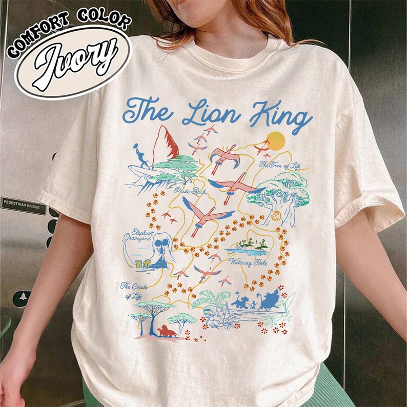May include: A white t-shirt with a colorful illustration of the map of Pride Rock from the movie The Lion King. The text "The Lion King" is printed in a bold font at the top of the shirt.