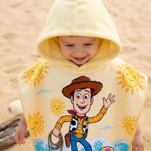 Puede incluir: Una toalla de playa con capucha amarilla con una imagen de dibujos animados de Woody de Toy Story. La toalla tiene un dise&ntilde;o de sol sonriente y una cuerda azul. El ni&ntilde;o lleva la toalla en una playa de arena.