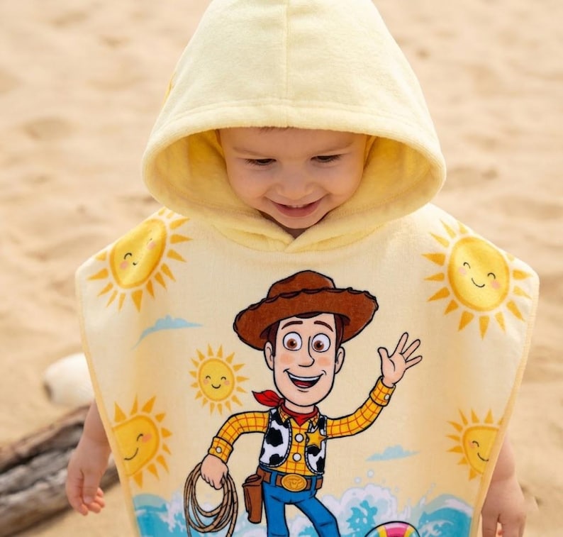 Puede incluir: Una toalla de playa con capucha amarilla con una imagen de dibujos animados de Woody de Toy Story. La toalla tiene un dise&ntilde;o de sol sonriente y una cuerda azul. El ni&ntilde;o lleva la toalla en una playa de arena.