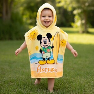 Puede incluir: Una toalla de playa con capucha amarilla con Mickey Mouse sosteniendo una tabla de surf. La toalla tiene un dise&ntilde;o de escena de playa con soles y olas, y el nombre "Anthon" est&aacute; impreso en la parte inferior. El ni&ntilde;o lleva la toalla y sonr&iacute;e.
