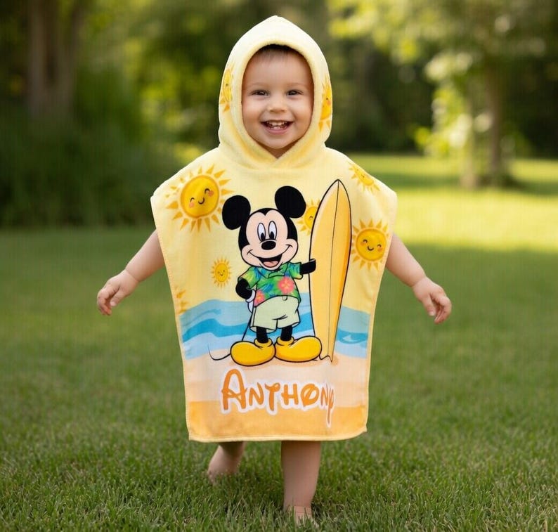 Puede incluir: Una toalla de playa con capucha amarilla con Mickey Mouse sosteniendo una tabla de surf. La toalla tiene un dise&ntilde;o de escena de playa con soles y olas, y el nombre "Anthon" est&aacute; impreso en la parte inferior. El ni&ntilde;o lleva la toalla y sonr&iacute;e.