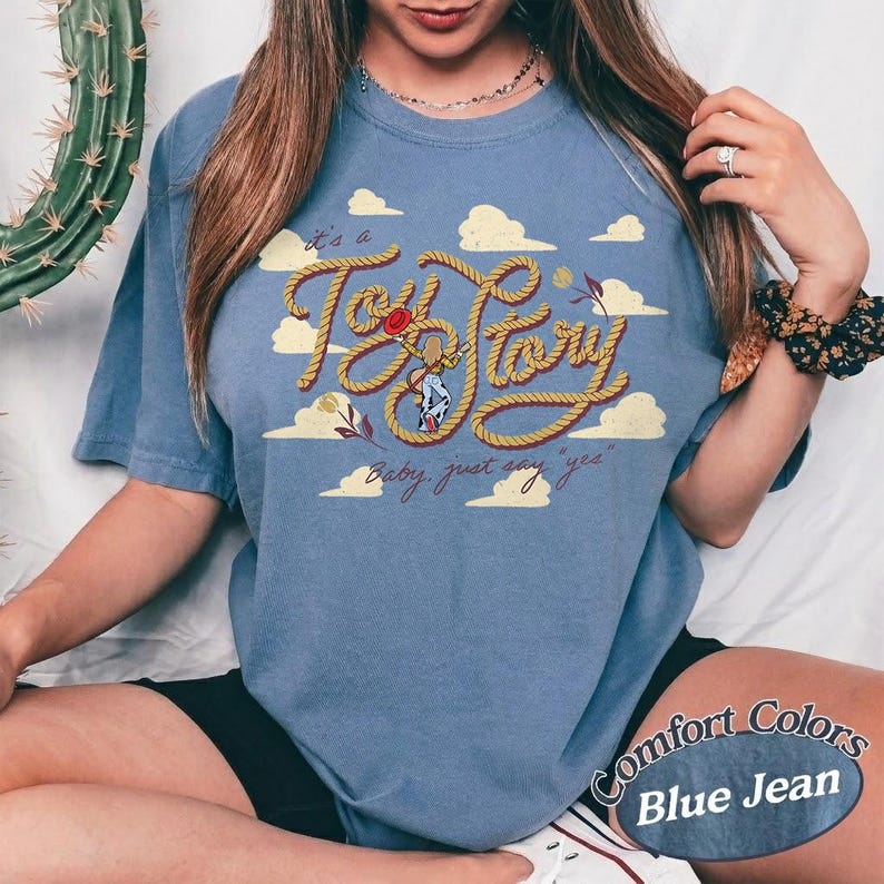 May include: A blue short-sleeve t-shirt featuring a graphic print of a rope shaped into the text "Toy Story" with a small cowboy figure in the center. The design includes yellow clouds and the cursive text "it's a" and "Baby, just say "yes"" in brown.