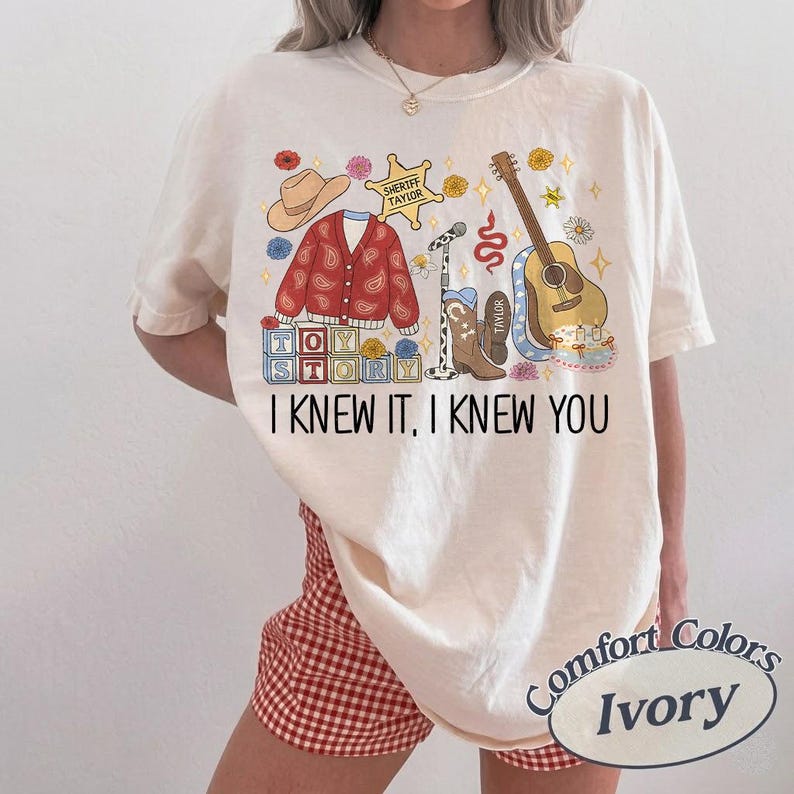 May include: An ivory t-shirt featuring a graphic print with a red paisley cardigan, a cowboy hat, a sheriff badge labeled Taylor, a microphone, a brown acoustic guitar, cowboy boots, and blocks spelling Toy Story with the text I KNEW IT, I KNEW YOU.