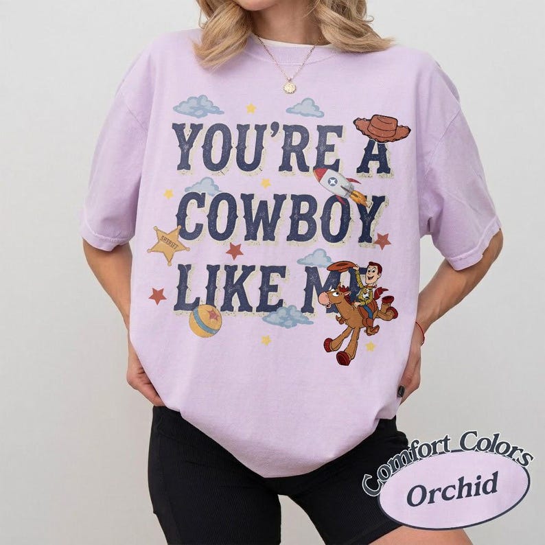 May include: An orchid-colored t-shirt featuring the dark blue text YOU'RE A COWBOY LIKE ME. The graphic includes a brown cowboy hat, a rocket ship, a yellow sheriff star, a colorful ball, and a cartoon character riding a brown horse.