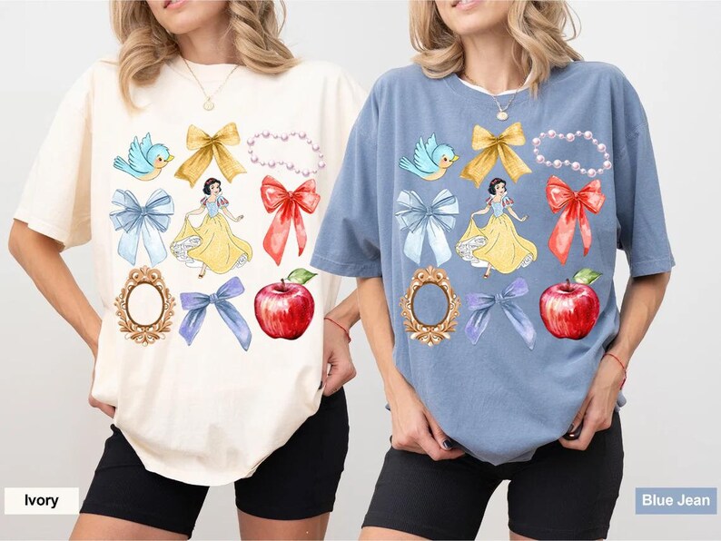 K&ouml;nnte beinhalten: Zwei T-Shirts mit einem Schneewittchen-Motiv. Das linke Shirt ist elfenbeinfarben mit einem blauen Vogel, einer roten Schleife und einem Spiegel. Das rechte Shirt ist jeansblau mit einem blauen Vogel, einer roten Schleife und einem Spiegel.