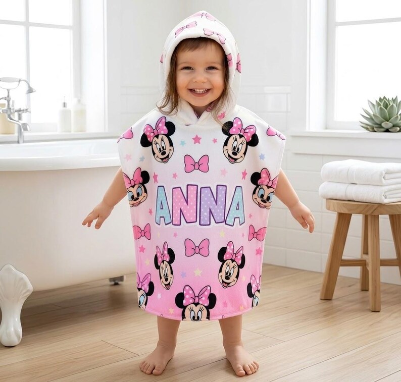 May include: A hooded, white and pink Minnie Mouse poncho towel with the name "ANNA" printed on the front. The towel features Minnie Mouse faces, pink bows, and stars. The child is standing in a bathroom.