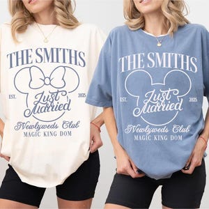 May include: Ivory and blue jean-colored t-shirts with the text "THE SMITHS" and "Just Married" in a Mickey Mouse-inspired design. The shirts also feature "Newlyweds Club" and "MAGIC KING DOM" text. The shirts are short-sleeved.