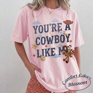 May include: A light pink short-sleeve t-shirt featuring the dark blue text "YOU'RE A COWBOY LIKE ME" printed on the front. The design includes illustrations of a cowboy hat, a rocket, a sheriff star, a ball, and a cowboy riding a horse.