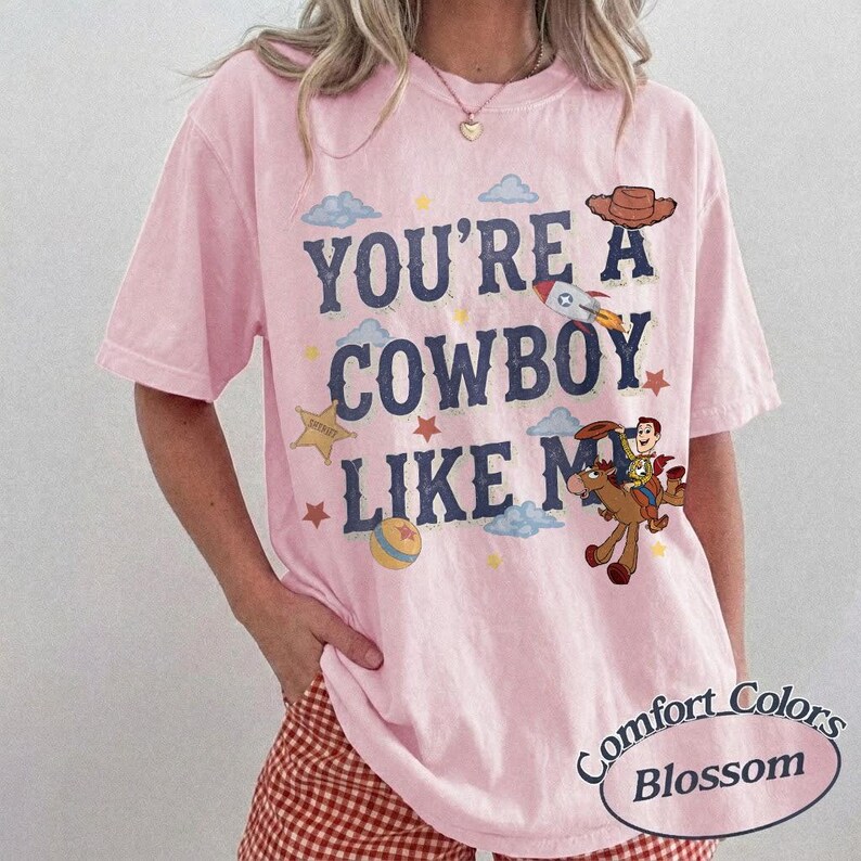 May include: A light pink short-sleeve t-shirt featuring the dark blue text "YOU'RE A COWBOY LIKE ME" printed on the front. The design includes illustrations of a cowboy hat, a rocket, a sheriff star, a ball, and a cowboy riding a horse.