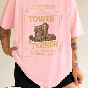 May include: Pale pink t-shirt with a gold-coloured graphic of the Hollywood Tower of Terror. The text on the shirt reads "Tower of Terror" and "You may not come back from the Twilight Zone." The shirt also has the text "That door is opening once again... and this time it's opening for you."