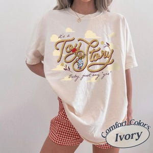 May include: An ivory t-shirt featuring a graphic design with the text "it's a Toy Story" written in a rope-style font, accompanied by the phrase "Baby, just say 'yes'" and illustrations of clouds, a red hat, and a small character figure.