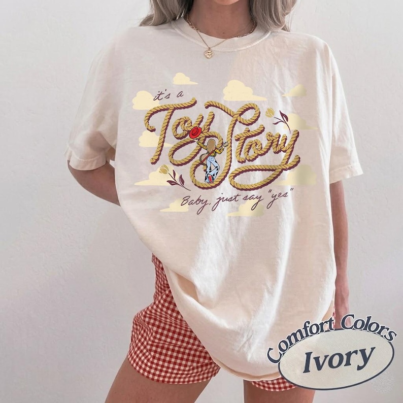 May include: An ivory t-shirt featuring a graphic design with the text "it's a Toy Story" written in a rope-style font, accompanied by the phrase "Baby, just say 'yes'" and illustrations of clouds, a red hat, and a small character figure.