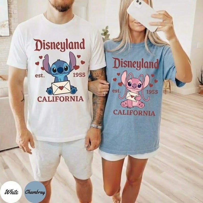 Op de afbeelding: Wit T-shirt met een Disneyland-afbeelding met een blauw stripfiguur dat een envelop vasthoudt. De tekst luidt "Disneyland est. 1955 CALIFORNIA". Het shirt wordt gecombineerd met een grijze korte broek.
