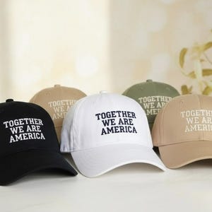 May include: Five baseball caps in black, white, beige, olive green, and tan. Each cap features the embroidered text "TOGETHER WE ARE AMERICA" in white. The caps are displayed on a white surface.