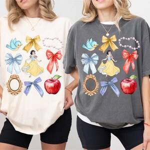 K&ouml;nnte beinhalten: Zwei T-Shirts mit einem Schneewittchen-Motiv. Die Shirts zeigen verschiedene Bilder, darunter Schneewittchen, V&ouml;gel, Schleifen, &Auml;pfel und Spiegel. Die Shirts sind in Elfenbein und Pfeffer erh&auml;ltlich.